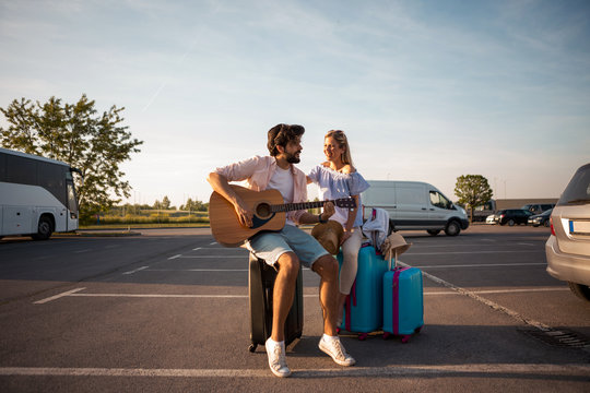 A handsome guy with a beard sings and plays on a guitar song for a beautiful blonde next to him while they are waiting for a bus for a tourist destination.
