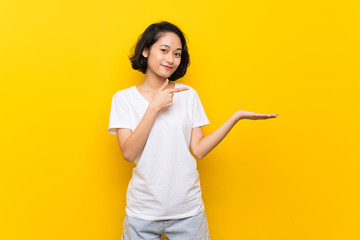 Asian young woman over isolated yellow wall holding copyspace imaginary on the palm to insert an ad - obrazy, fototapety, plakaty
