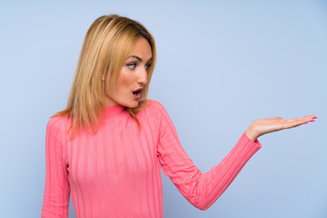 Fototapeta premium Young blonde woman with pink sweater over isolated blue background holding copyspace imaginary on the palm