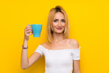 Young blonde woman over isolated yellow wall holding hot cup of coffee