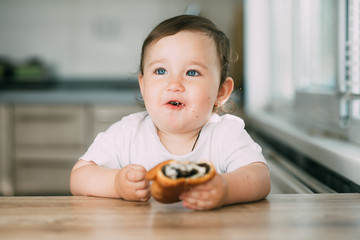 a little girl in the kitchen during the day eating a bun with poppy seeds is very appetizing