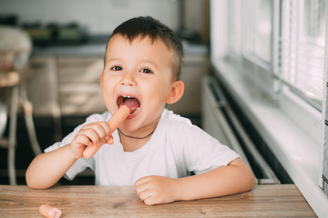 Portrait of a adorable little boy in a white t-shirt at the table eating a sausage with his hands