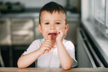 Portrait of a adorable little boy in a white t-shirt at the table eating a sausage with his hands