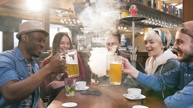 Company of hipster young people chatting, clinking with glass of beer and smoking shisha or hookah in bar.