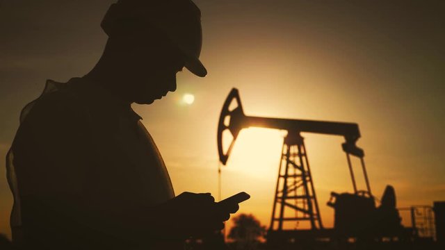 Silhouette of man engineer with phone overseeing the site of crude oil production at sunset.