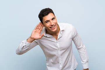 Handsome man over blue wall listening to something by putting hand on the ear