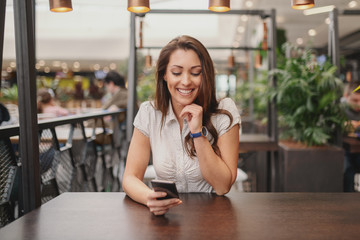 Portrait of beautiful Caucasian brunette sitting in cafe and reading message on smart phone.