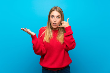 Woman with red sweater over blue wall making phone gesture and doubting