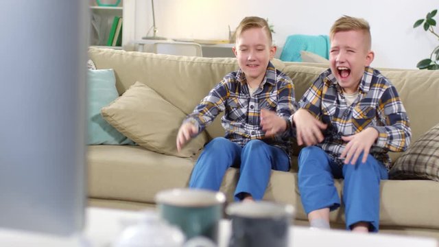 Full shot of 9-year-old Caucasian identical twin brothers sitting in living room at home, watching something funny on TV, pointing towards screen and rolling around on couch with hysterical laughter