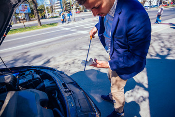 A businessman has parked his car at the side of the boulevard while he checks his car oil, on a sunny busy day.