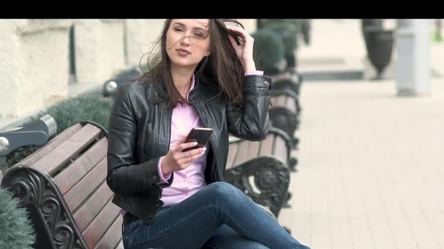 an attractive girl in a leather black jacket sits on a bench and looks at the phone. He raises his head and smiles at the camera. The strong wind ruffled her hair.