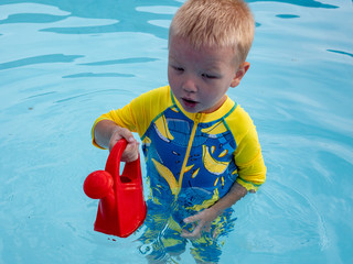 Little blonde boy  in hot summer day swim in carcas pool  in swimwear for boys. Kid is holding in hand red watering can