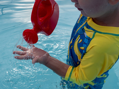 Little blonde boy  in hot summer day swim in carcas pool  in swimwear for boys. Kid is holding in hand red watering can