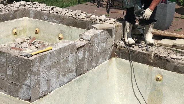 Construction worker demolishing coping, tile and concrete around empty swimming pool and hot tub for remodel