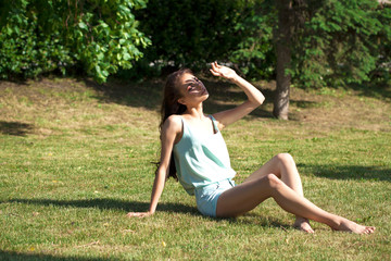 Fototapeta premium Young brunette woman in a turquoise blouse posing sitting on a green lawn in a summer park
