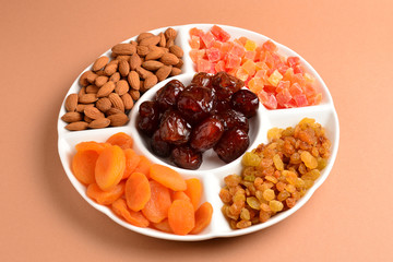 Mix of dried fruits and nuts on a white plate. Apricot, almond, raisin, dates fruit. On a brown background. Space for text or design.