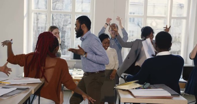 Happy young African businessman doing fun cheerful holiday celebration dance with colleagues at healthy workplace.