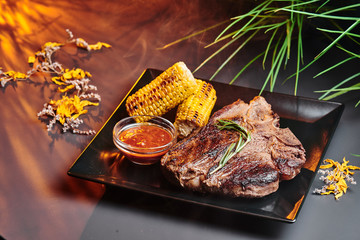 Juicy T-Bone steak with grilled corn and barbecue sauce on a black square plate on a dark smoky...