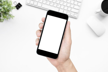 Hand holding smartphone with blank mock up screen over white office desk table. Top view image.