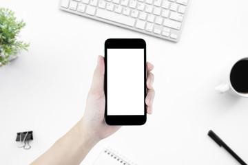 Hand holding smartphone with blank mock up screen over white office desk table. Top view image.