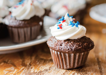 Mini cupcakes decorated in red, white, and blue sprinkles.  Blurred additional cupcakes in background.  Wooden table.