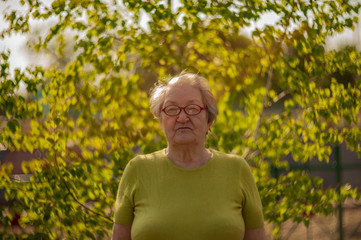 Elderly woman in yellow T-shirt and glasses posing in front of camera against background of bright green birch leaves