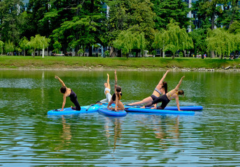 Yoga on sup board. Young girls paddling on SUP board on the lake at city. Group women is practicing (doing) yoga, fitness, pilates and meditation on a SUP board. Awesome active training in outdoor. 