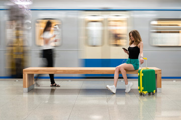 Happy girl traveler waiting for train at metro station sitting on stone bench with legs stretched out on suitcase