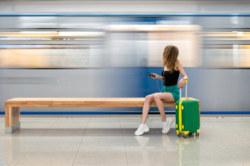 a young beautiful european girl with a small green suitcase is sitting on a bench at the railway station waiting for the train. Theme travel alone