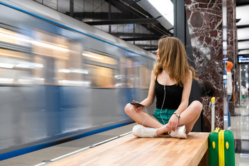 European girl sitting on a bench with a phone in her hands and in the headphones, next to a travel suitcase. It is located at the railway station or subway station. Theme of independent travel.
