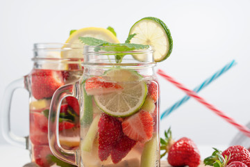 Two mugs of fruit refreshing water made from strawberries, lemon, mint and lime in the background, drinking straws and fresh strawberries.