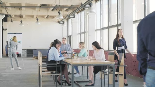 Business team holds a meeting at a large table. An employee gets up and goes to colleagues for a glass presentation board. Coworking. Loft style office space