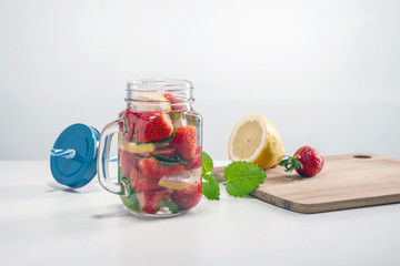 Mug of fruit water with strawberries, mint and lemon, stand on a wooden stand next to fresh strawberries, mint leaf, sliced lemon and a cap with a straw for drinking.