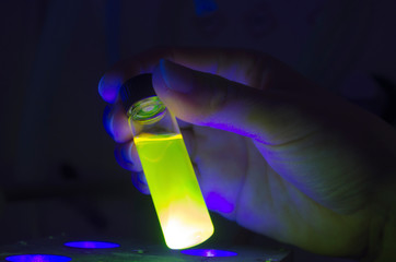 Researcher holding green photochemical reaction in glass vial under UV light in a dark chemistry laboratory