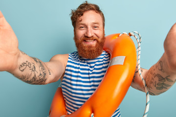 Joyful redhead man being on holiday at sea, makes selfie, poses against blue background with...