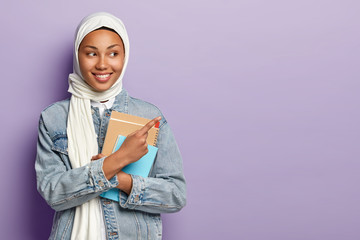 Isolated shot of satisfied Muslim student shows awesome advert, points at upper right corner, wears white veil and jean jacket, carries notepads , prepares for classes, isolated over purple wall