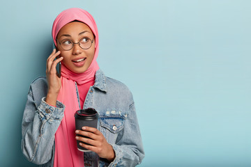 Portrait of delighted Arabian woman makes arrangement via cell phone, holds paper cup of coffee, receives good offer, looks aside, dressed in denim jacket, has thoughtful expression, isolated on blue