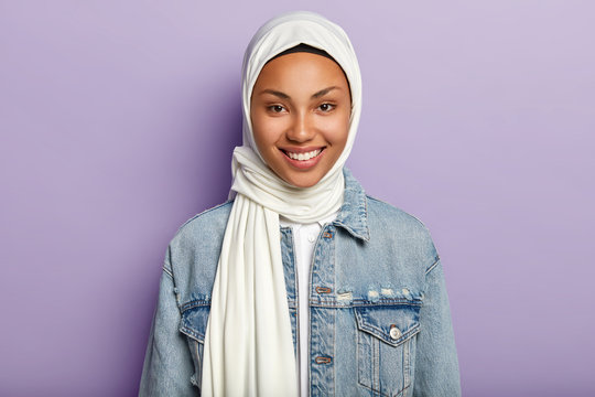 Portrait Of Joyful Good Looking Woman Has Islamic Views, Smiles Gently At Camera, Glad To Hear Good News, Covers Head With White Veil, Models Against Purple Background. Positive Emotions And Feelings