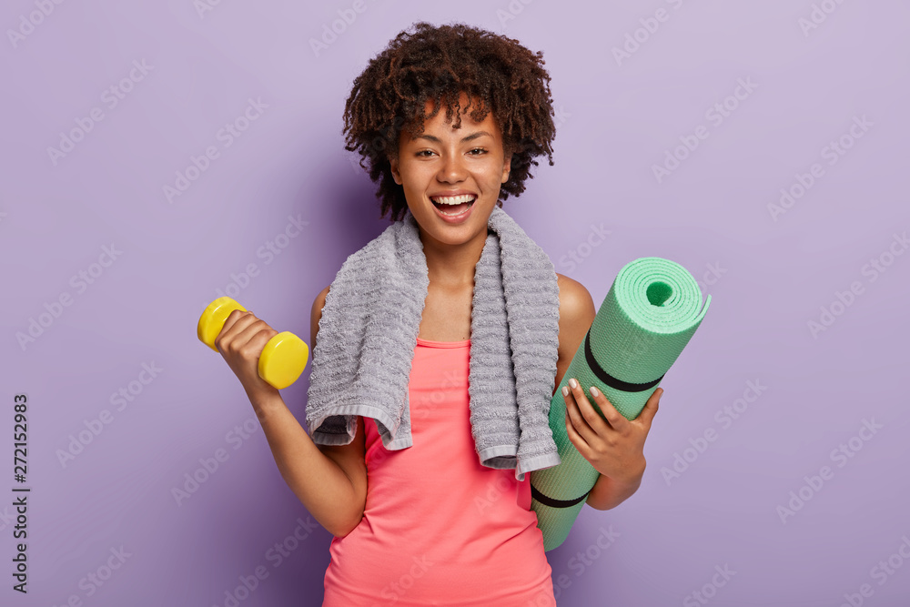 Wall mural happy afro american woman lifts small weights, holds green karemat, ready for yoga exercises, smiles