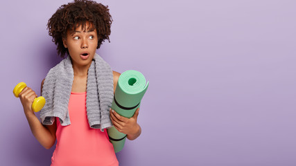 Studio shot of surprised Afro woman does exercises with dumbbell, trains endurance, uses fitness mat, dressed in sporting clothes, focused aside with astonishment, models over purple wall, blank space