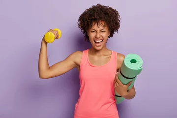 Gordijnen Funny happy dark skinned woman raises hand with dumbbell, shows biceps, holds rolled fitness mat, smiles broadly, wears pink casual vest, isolated over purple background. Fitness training at home © Wayhome Studio
