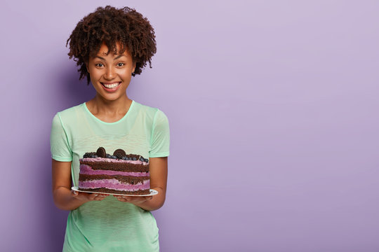 Happy Afro American woman holds delicious birthday cake with blueberry, treats guests with tasty sweet dessert, dressed in casual green t shirt, stands in studio against purple background, free space