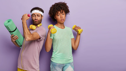 Tired active multiethnic woman and man stand closely, train biceps with dubbells, wear t shirt, use karemat for yoga training, involved in sport, isolated over purple wall. Together we achieve more