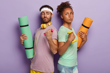 Diverse sporty couple stand backs, hold dumbbells, fitness mats, has confident determined facial expressions, use sport equipment, listen music in headphones, stand over purple wall. Athletic family