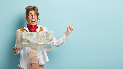 Positive female traveler holds paper map, shows direction, being lost in unknown place, wears round spectacles, red bandana, points at free space, has glad look, isolated over blue background - obrazy, fototapety, plakaty