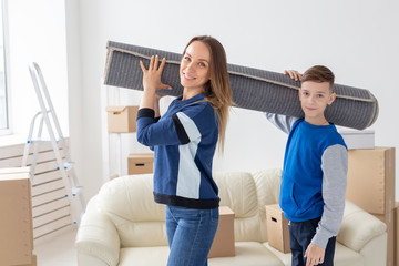 Smiling caucasian mother and a charming son are holding a folded carpet on their shoulders in a new living room intending to spread it in a new apartment