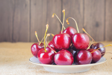 Ripe fresh sweet cherry on a small white plate. Behind - a wooden wall. The concept of vitamins, healthy nutrition, vegetarianism, ripening berries and fruits. Cozy still life, minimalism.