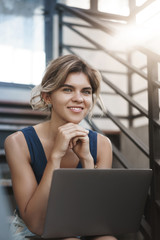 Fototapeta premium Vertical close-up shot attractive happy smiling blond european young woman sit stairs outdoor hold laptop knees working freelance, studying, gather information before classes research via internet