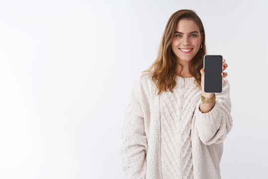 Girl wanna hear your opinion showing you smartphone display. Attractive charming friendly-looking woman extending hand holding mobile phone sharing funny picture smiling broadly white background