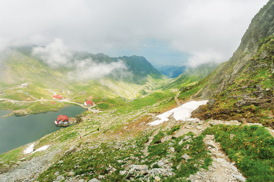 lake balea in dramatic weather. beautiful destination in fagaras mountain of romania. view through clouds from the top of a ridge. steep slopes with rocks among the grass. famous road 7C in the valley